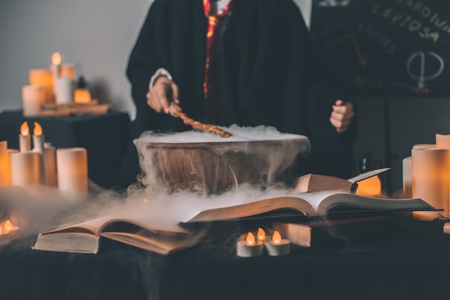 A person holding a wand over a smoky cauldron, with open books lying around.