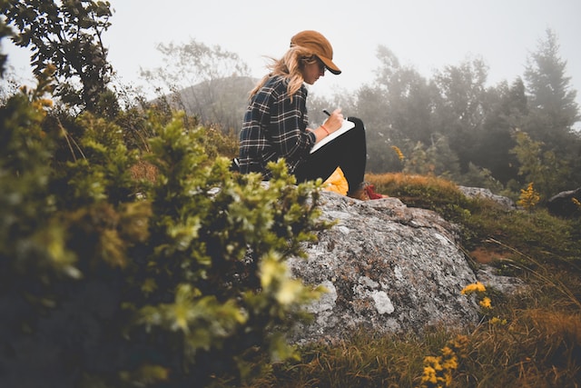 A woman sitting on a rocky hill, writing on a notepad.