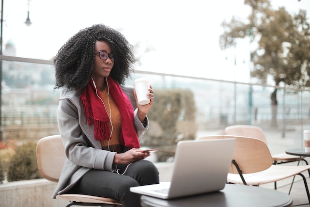 A woman holding a smartphone and a drink while looking at an open laptop.