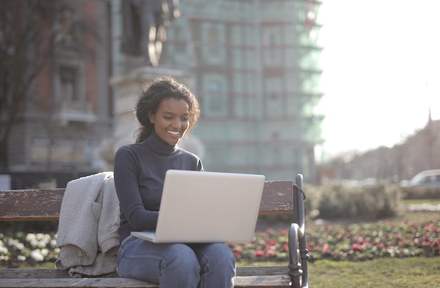 A smiling woman, sitting on a bench outside, while working on her laptop.