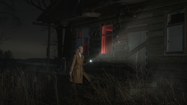 A lady in a brown overcoat with a flashlight in her hand walking outside an eerie-looking house at night