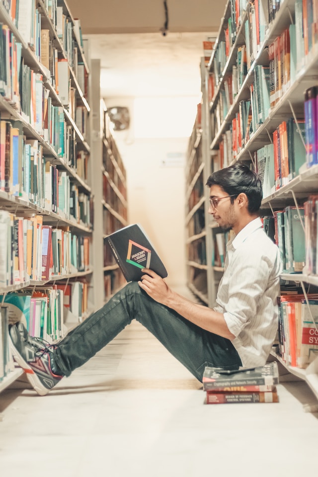 A guy in glasses sitting on the floor with an open book on his knees