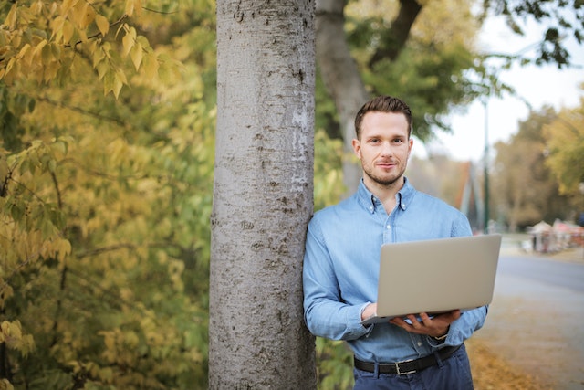 Guy in blue shirt holding a laptop and leaning against a tree