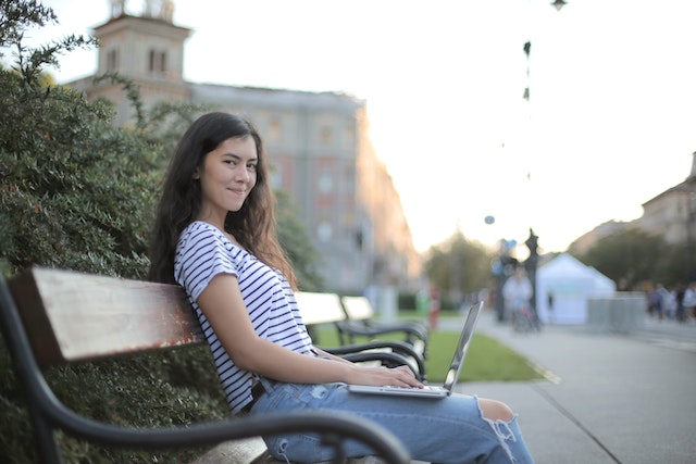 Young woman in striped shirt sitting on a bench with laptop on her lap