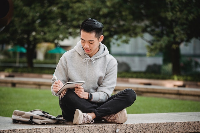 A guy in a hoodie sitting cross-legged while writing outside